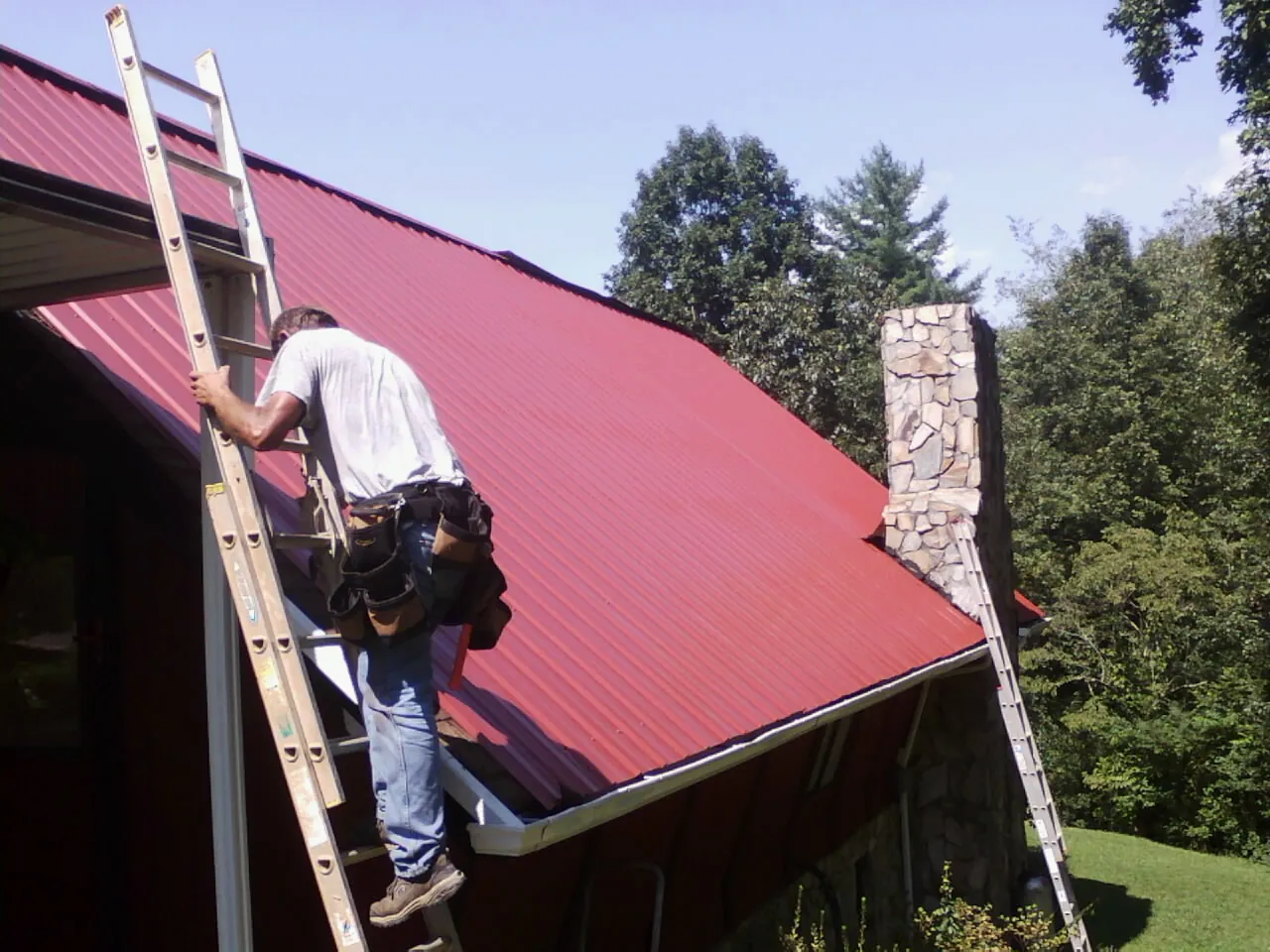 Roofer on a ladder beside a red metal roof and stone chimney — Soffit & Fascia Repair by Roofing Brothers in Hockley